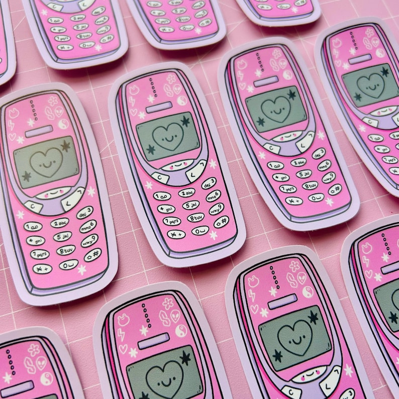 May include: Pink retro cell phone stickers with a heart-shaped smiley face on the screen. The stickers have a white border and are decorated with stars, flowers, and other small designs. The phone buttons are labeled with numbers and symbols.