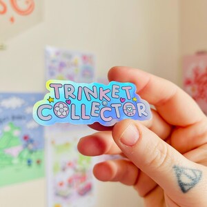 May include: A holographic sticker with the words "TRINKET COLLECTOR" in a bubble font. The sticker has a flower, hearts, and a retro video game console graphic. The sticker is held in a hand.