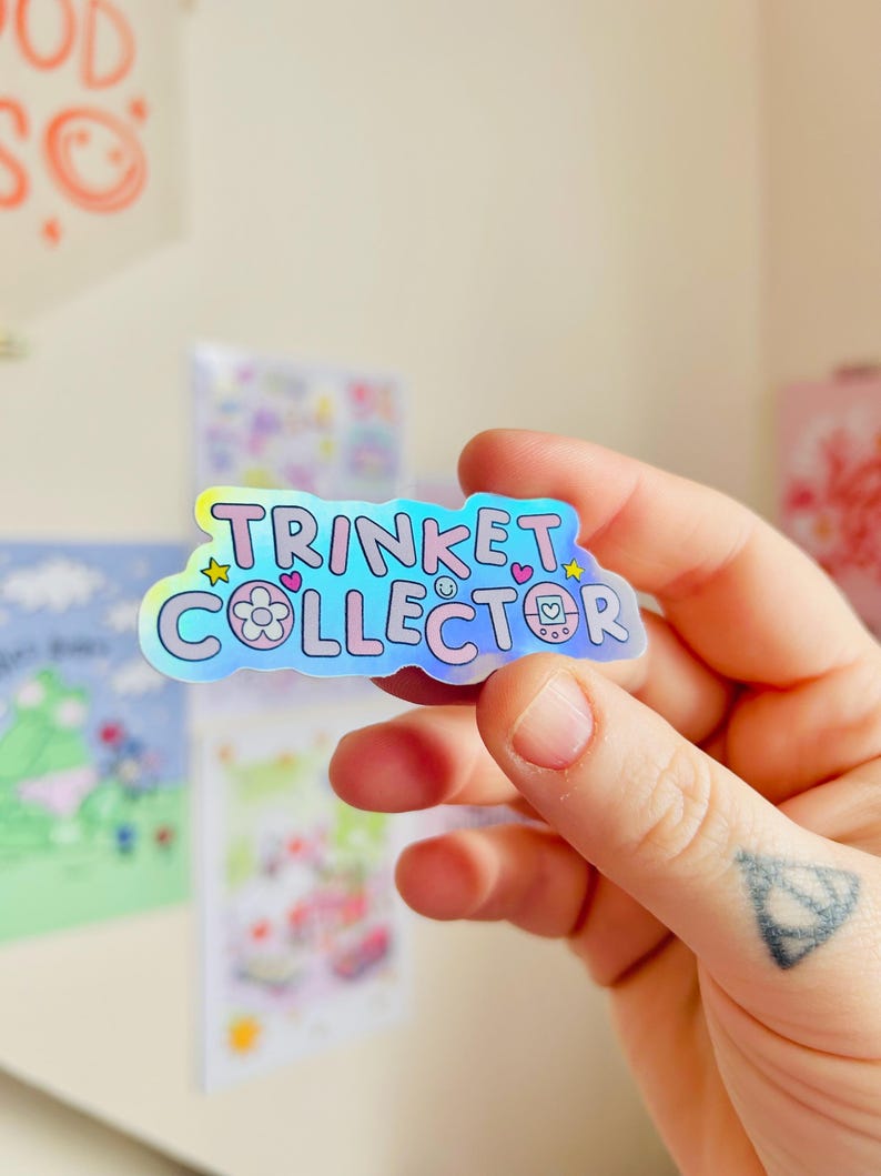 May include: A holographic sticker with the words "TRINKET COLLECTOR" in a bubble font. The sticker has a flower, hearts, and a retro video game console graphic. The sticker is held in a hand.