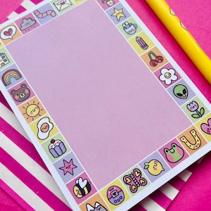 May include: A rectangular sheet of light purple stationery paper with a border of small, colorful, cartoon-style icons including a rainbow, bear, sun, egg, cupcake, bee, mountain, butterfly, frog, flower, alien, and mushroom, paired with a yellow pen.