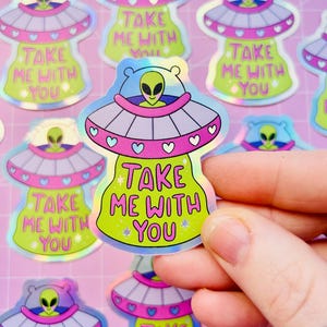 May include: Holographic stickers featuring a cartoon UFO with an alien pilot. The UFOs are pink, purple, and green, with the text "TAKE ME WITH YOU". The stickers have a shiny, iridescent finish and are scattered on a pink background.