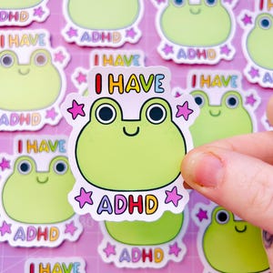 Può includere: Una collezione di adesivi a forma di rana verde con la scritta "I HAVE ADHD". Gli adesivi raffigurano un simpatico ranocchio con grandi occhi e una bocca sorridente, circondato da stelle rosa. Il testo è multicolore.