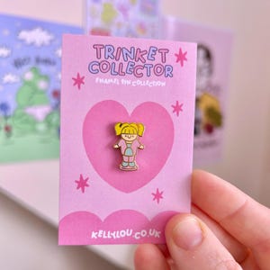 May include: A pink enamel pin of a cartoon girl with yellow pigtails, on a pink card with a pink heart. The card reads "TRINKET COLLECTOR" and "ENAMEL PIN COLLECTION". The website "KELLYLOU.CO.UK" is printed at the bottom of the card.