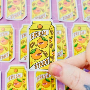 Könnte beinhalten: Ein Bogen mit Aufklebern, die Cartoon-Orangensaftkartons darstellen. Die Aufkleber sind gelb mit Orangenillustrationen, dem Text "FRESH" und "START". Ein Aufkleber wird von einer Hand gehalten.