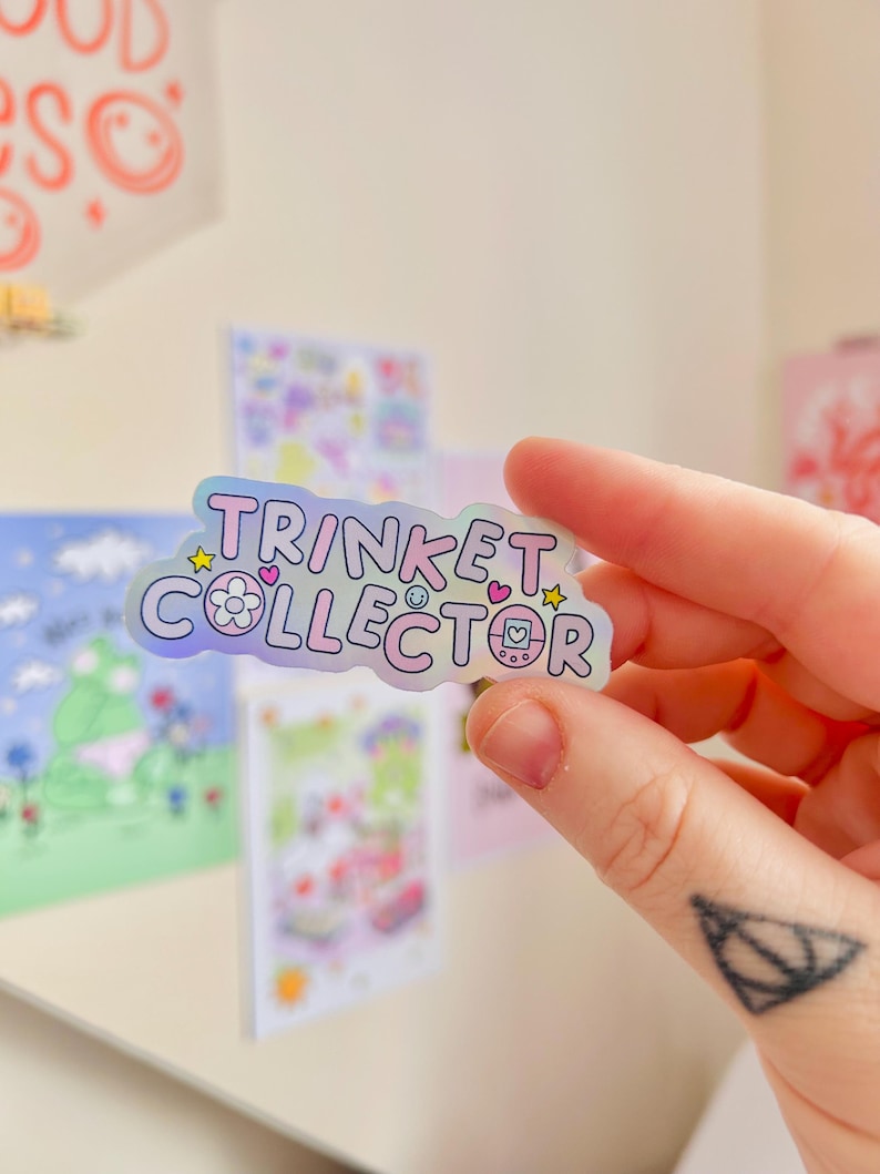 May include: A holographic sticker with the words "TRINKET COLLECTOR" in a bubble font. The sticker has a pastel color scheme with stars, hearts, and flower icons. The sticker is held by a person's hand.