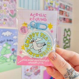 Könnte beinhalten: Transparenter Acryl-Schlüsselanhänger mit einem Cartoon-Möwen-Design. Der Schlüsselanhänger hat einen hellblauen Verschluss und einen kleinen kreisförmigen Anhänger. Der Text auf dem Schlüsselanhänger lautet "CHIPPY TEA ENTHUSIAST" und "Kelly Lou". Zusätzlicher Text unten lautet "FIND MORE ILLUSTRATED AT KELLYLOU.CO.UK".