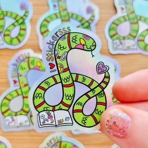 May include: A holographic sticker featuring a green snake with red stripes, a pink heart on its tail, and a small open book at the base. The snake has a smiling face and the text I love stickers is printed in black along the curved body of the snake.