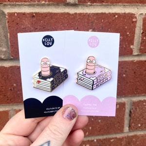 May include: Two enamel pins featuring a pink worm with glasses emerging from a stack of books. The text on the pins reads "Book Worm". The pins are on white cards with the text "Kelly Lou" and website information.