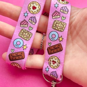 May include: A pink fabric lanyard featuring a repeating pattern of cartoon-style sweets, including brown rectangular Bourbon biscuits, round cookies with red hearts, blue donuts, and purple swirls, accented with small yellow stars and green hearts.