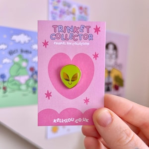 May include: A green alien enamel pin with gold detailing, presented on a pink card. The card features the text "Trinket Collector" and "Enamel Pin Collection", along with a heart and star motifs. The website "kellylou.co.uk" is printed at the bottom.