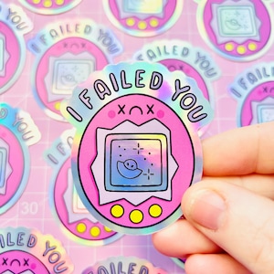 Pode incluir: Autocolantes holográficos com um design rosa retro de animal de estimação digital e o texto "I FAILED YOU". Os autocolantes têm uma estética de desenho animado, com um pequeno ecrã que mostra uma lua e estrelas. Os autocolantes estão espalhados numa superfície rosa.