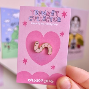 May include: A pink enamel pin in the shape of a worm, with gold-colored outlines, is displayed on a pink card. The card has the text "TRINKET COLLECTOR" and "ENAMEL PIN COLLECTION". The card also features a heart and star designs.