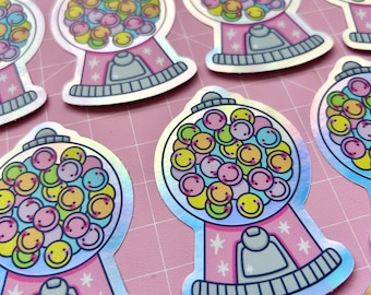 May include: A collection of holographic stickers featuring a gumball machine design. The stickers have a pink base with a silver top and base. The gumball machine is filled with colorful gumballs with smiley faces.