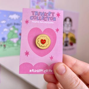 May include: A pink card featuring a gold-coloured enamel pin with a red heart design. The pin is flower-shaped. The card reads "TRINKET COLLECTOR ENAMEL PIN COLLECTION" and "KELLY LOU.CO.UK". The background includes other cards.