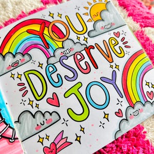 May include: A colourful illustration featuring rainbows, clouds and stars. The text "You Deserve Joy" is written in a playful, hand-drawn style.