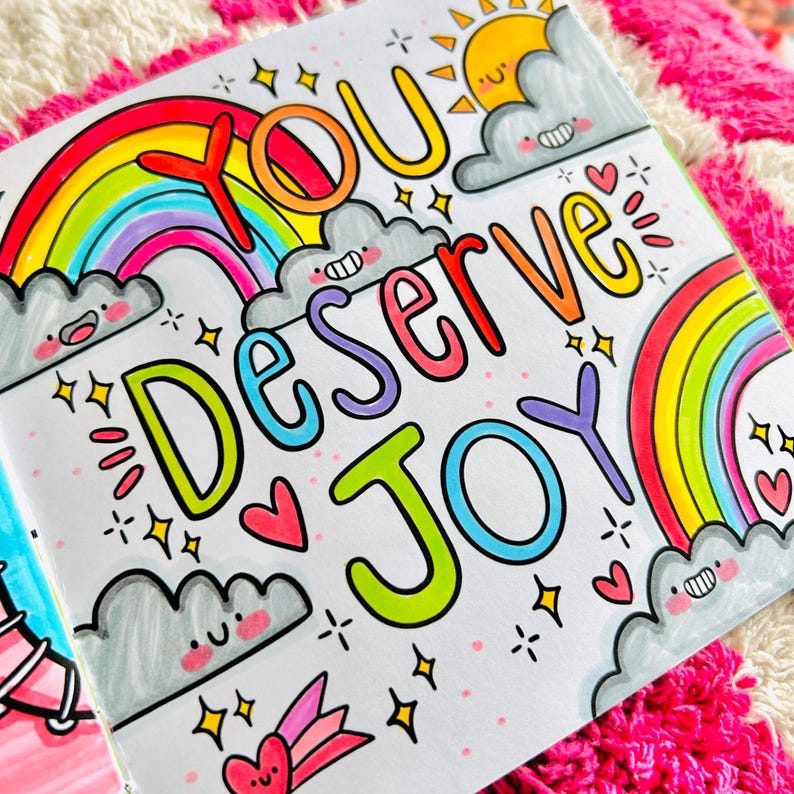 May include: A colourful illustration featuring rainbows, clouds and stars. The text "You Deserve Joy" is written in a playful, hand-drawn style.
