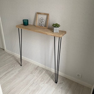 Op de afbeelding: Een houten consoletafel met zwarte haarspeldpoten. De tafel heeft een lichte houtkleur en is versierd met een klein ingelijst printje van een bloem en een potplant.