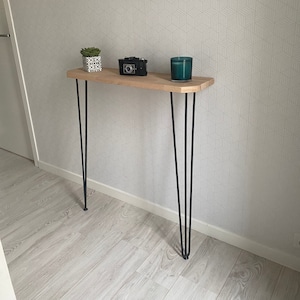 Op de afbeelding: Een houten consoletafel met zwarte haarspeldpoten. De tafel heeft een lichte houtkleur en is versierd met een kleine plant in een pot, een vintage camera en een teal kaars.