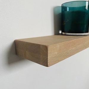 May include: A light brown wooden shelf with a teal glass candle on top. The shelf is mounted to a white wall.