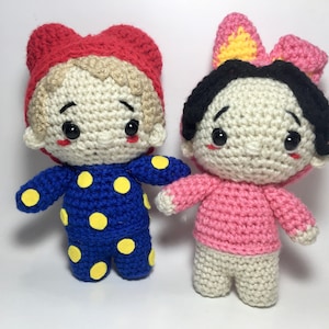 Può includere: Due bambole all'uncinetto fatte a mano. Una bambola indossa un cappello rosso e un pigiama blu con pois gialli. L'altra bambola indossa un maglione rosa e ha un fiocco rosa tra i capelli. Entrambe le bambole hanno occhi neri e guance rosee.