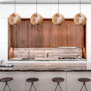 May include: A modern kitchen island with a brown granite countertop and four wooden bar stools. The island is situated in front of a white kitchen with brown cabinets and a backsplash of light brown stone. Four round, woven pendant lights hang above the cabinets.