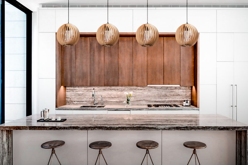 May include: A modern kitchen island with a brown granite countertop and four wooden bar stools. The island is situated in front of a white kitchen with brown cabinets and a backsplash of light brown stone. Four round, woven pendant lights hang above the cabinets.
