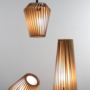 Pu&ograve; includere: Tre lampade in legno con paralumi a lamelle. Le lampade sono accese e proiettano una luce calda. La lampada superiore &egrave; appesa al soffitto, mentre le altre due sono su un tavolo.