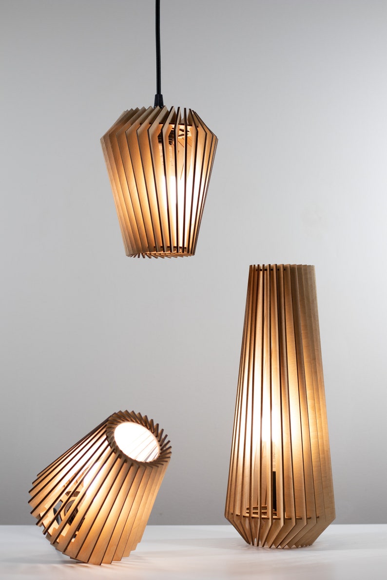 Pu&ograve; includere: Tre lampade in legno con paralumi a lamelle. Le lampade sono accese e proiettano una luce calda. La lampada superiore &egrave; appesa al soffitto, mentre le altre due sono su un tavolo.