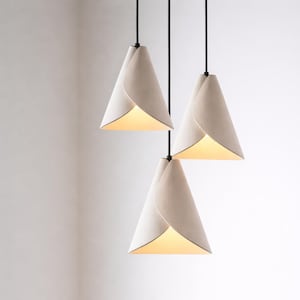 A cluster of three handcrafted white pendant lights hangs at staggered heights, featuring soft sculptural folds and a warm ambient glow against a clean, minimalist background.