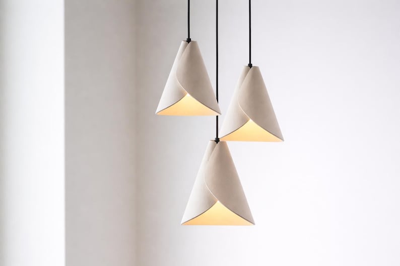 A cluster of three handcrafted white pendant lights hangs at staggered heights, featuring soft sculptural folds and a warm ambient glow against a clean, minimalist background.