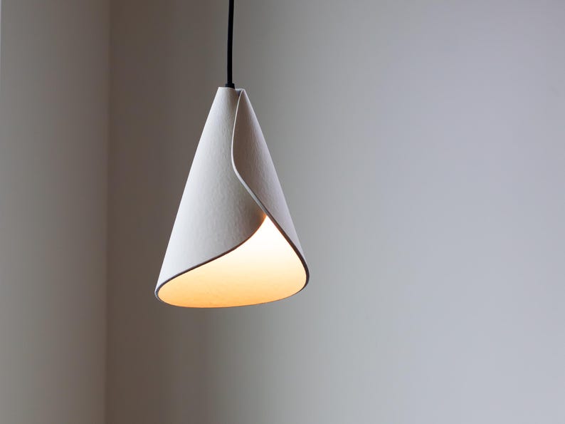 Handcrafted white 3D-printed pendant light with a twisted cone form, glowing warmly against a minimalist gray wall.