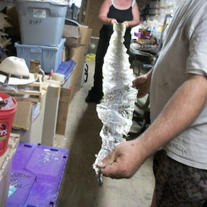May include: A person holding a long, textured, silver-colored object, possibly a hide or skin, in a cluttered workshop setting. The object has a ridged pattern and is held vertically. Other items include boxes, a hat, and a coffee can.