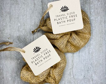 Puede incluir: Dos esponjas de baño de viaje sin plástico hechas de un material tejido de color natural. Cada esponja tiene una etiqueta adjunta con hilo, que muestra el texto "TRAVEL SIZED PLASTIC FREE BATH POUF" y "All Natural - Biodegradable".