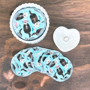 May include: A stack of round, reusable facial rounds with an otter print in a white, scalloped-edge dish. Additional rounds are scattered nearby. A small, white heart-shaped dish is also present. The rounds feature a light blue background.