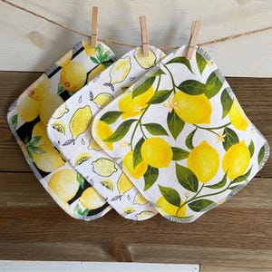 May include: Three square cloths with lemon patterns hang from a string with wooden clothespins. The cloths feature bright yellow lemons and green leaves on white and black backgrounds. The cloths are made of a soft, absorbent material.