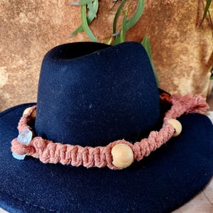 May include: A navy blue fedora hat with a decorative, woven band in a muted orange colour. The band is adorned with wooden beads and a small, clear crystal. The hat is a stylish accessory.