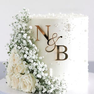 Könnte beinhalten: Eine weiß glasierte Torte, verziert mit weißen Rosen, Schleierkraut und goldfarbenen Initialen "N & B". Der Kuchen ist mit kleinen weißen, perlenartigen Streuseln verziert. Ein wunderschöner Kuchen für eine Hochzeit oder einen besonderen Anlass.