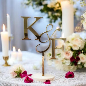 Könnte beinhalten: Ein goldfarbener Tortenaufsatz für Hochzeiten mit den Buchstaben "K & F". Die Buchstaben sind mit einem dekorativen Et-Zeichen verbunden und auf einem weißen Sockel montiert. Der Aufsatz ist von Kerzen und Blumen umgeben.