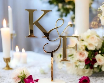 May include: A gold-colored wedding cake topper featuring the letters "K & F". The letters are connected with a decorative ampersand and mounted on a white base. The topper is surrounded by candles and flowers.