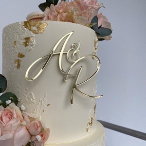 Könnte beinhalten: Eine zweistöckige Hochzeitstorte mit weißem Zuckerguss, floralen Akzenten und Goldblattdetails. Ein goldfarbenes Monogramm mit den Buchstaben "A&R" sitzt auf der Torte. Rosa Rosen und Grünzeug schmücken die Basis und die Oberseite der Torte.