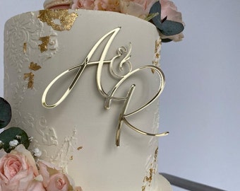 May include: A two-tiered wedding cake with white frosting, floral accents, and gold leaf details. A gold-colored monogram with the letters "A&R" sits atop the cake. Pink roses and greenery decorate the base and top of the cake.
