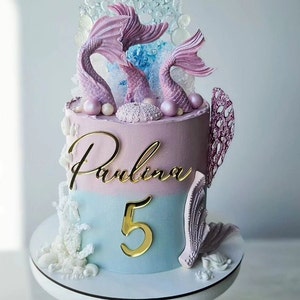 Könnte beinhalten: Eine zweistöckige Torte, die mit einem Meerjungfrauen-Motiv verziert ist. Die untere Etage ist hellblau mit einer goldenen Zahl "5" darauf. Die obere Etage ist hellrosa mit dem Namen "Paulina" in Gold geschrieben. Die Torte ist mit weißem Korallen, rosa und lila Meerjungfrauen und perlmuttartigen Dekorationen verziert.