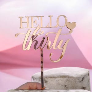 Könnte beinhalten: Ein goldfarbener Tortenaufsatz mit den Worten "HELLO thirty" in einer Schreibschrift. Ein kleines Herz befindet sich neben dem Wort "HELLO". Der Aufsatz wird von einer Hand gehalten und vor einem rosa Hintergrund platziert.