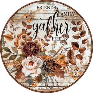 May include: A round wooden sign with a distressed white wood grain background. The text reads FRIENDS & FAMILY gather HERE in black script and serif fonts. An arrangement of brown autumn leaves, a dark red flower, a white flower, and a pinecone.