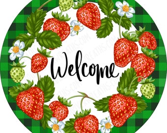 May include: A round sign featuring a green and black buffalo plaid border and a white center. The design includes a wreath of red strawberries, green leaves, and white flowers surrounding the black cursive text "Welcome" in the middle.