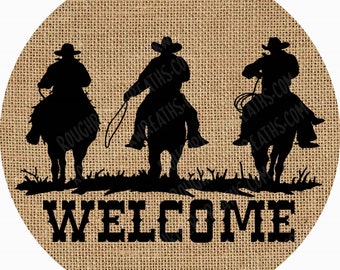 May include: A circular sign with a tan burlap texture background featuring a black silhouette of three cowboys on horseback. The cowboys are positioned above the word WELCOME printed in a bold, black, western-style serif font.