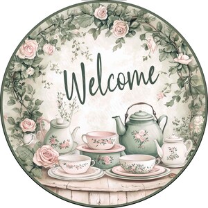 May include: A round sign featuring the word Welcome in dark green script, surrounded by a border of pink roses and green foliage. The scene includes a green teapot, a white teapot, and several teacups with floral patterns on a wooden table.