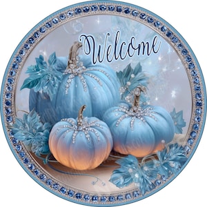 May include: A circular sign featuring three light blue pumpkins with orange-tinted bases, adorned with sparkling crystal accents on the stems and ribs. The word Welcome is written in script above the pumpkins, framed by a border of blue rhinestones.