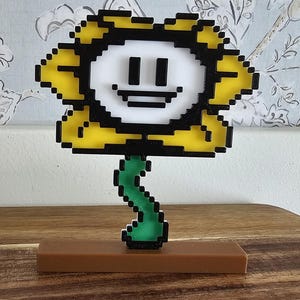 May include: A pixelated flower figurine with a yellow and black petal design, a white face, and a green stem. The figurine is mounted on a brown rectangular base, set against a floral wallpaper background.