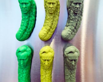 May include: A set of six fridge magnets shaped like pickles, each featuring a sculpted human face. The magnets are arranged in two rows and three columns, displaying three distinct colours: vibrant green, bright yellow, and muted olive green.